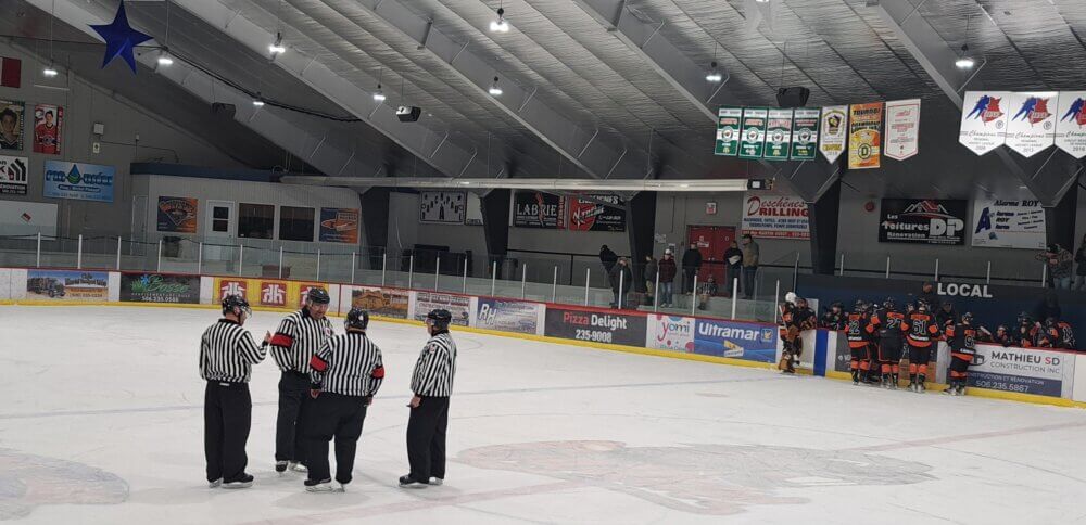 Quatre officiels discutent sur le premier tiers d'une patinoire intérieur. À droite, les joueurs des castors de Saint-Quentin sont à leur banc.