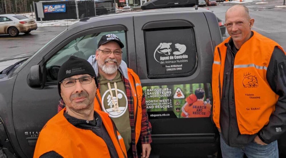 Trois hommes portant une veste orange devant une camionnette grise, lors d'une journée d'hiver pluvieuse.