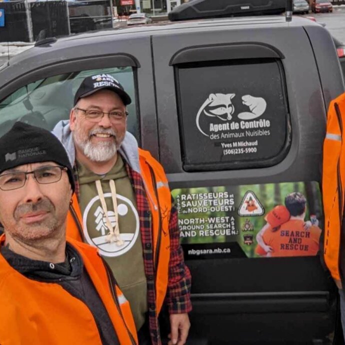 Trois hommes portant une veste orange devant une camionnette grise, lors d'une journée d'hiver pluvieuse.