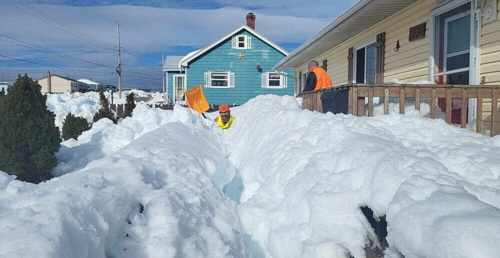 Un homme et son coéquipier sont enserrés d’une quantité importante de neige devant le balcon d’une résidence. Vêtu d’un dossard orange, les deux hommes sont en train de pelleter.