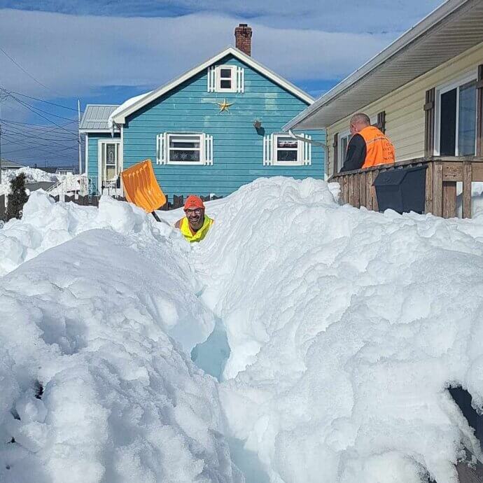 Un homme et son coéquipier sont enserrés d’une quantité importante de neige devant le balcon d’une résidence. Vêtu d’un dossard orange, les deux hommes sont en train de pelleter.