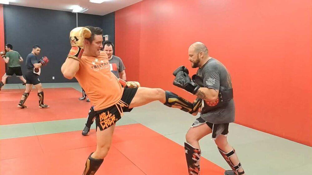 Un entraineur de boxe thaïlandaise donne un coup de pied à son élève, un homme d’une grande carrure, au niveau du ventre. L’élève porte un coussin protecteur.