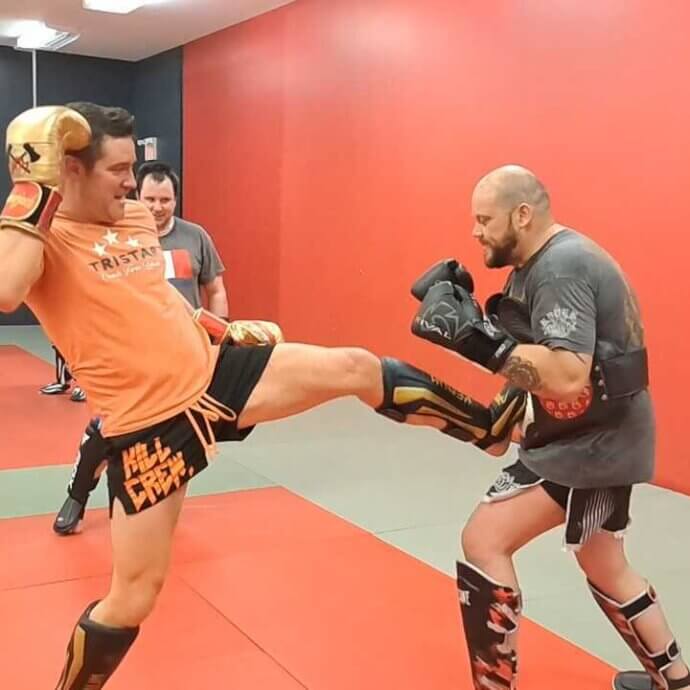 Un entraineur de boxe thaïlandaise donne un coup de pied à son élève, un homme d’une grande carrure, au niveau du ventre. L’élève porte un coussin protecteur.