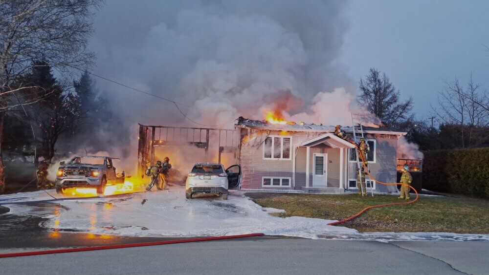 Maison blanche en flamme, tard en soirée. Il y a des pompiers sur le toit et un véhicule est également en flamme devant la maison.