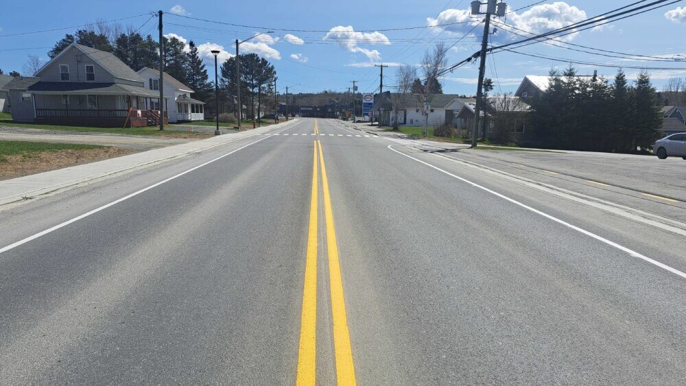 La rue Notre-Dame à Kedgwick. Photo prise en pleine centre de la ligne double jaune sur une rue asphaltée neuve, lors d'une journée de mai ensoleillée.