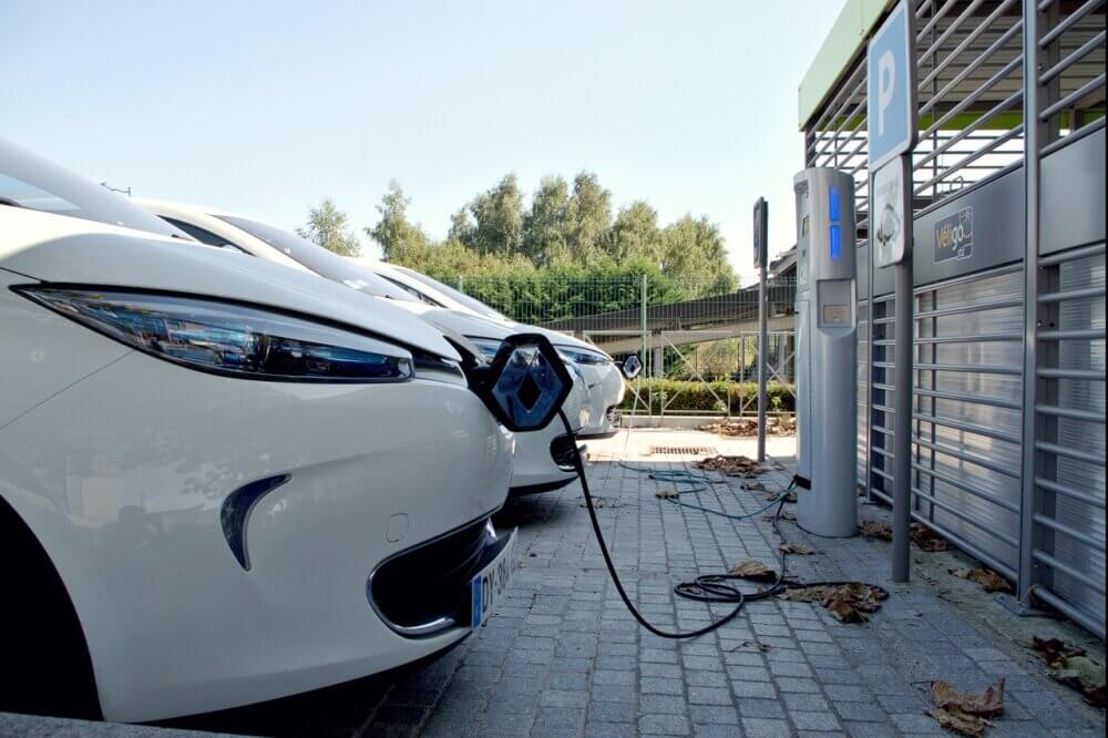 Place de stationnement. On aperçoit le devant d'un véhicule blanc électrique en plein chargement devant une borne de recharge, lors d'une journée d'été ensoleillée.