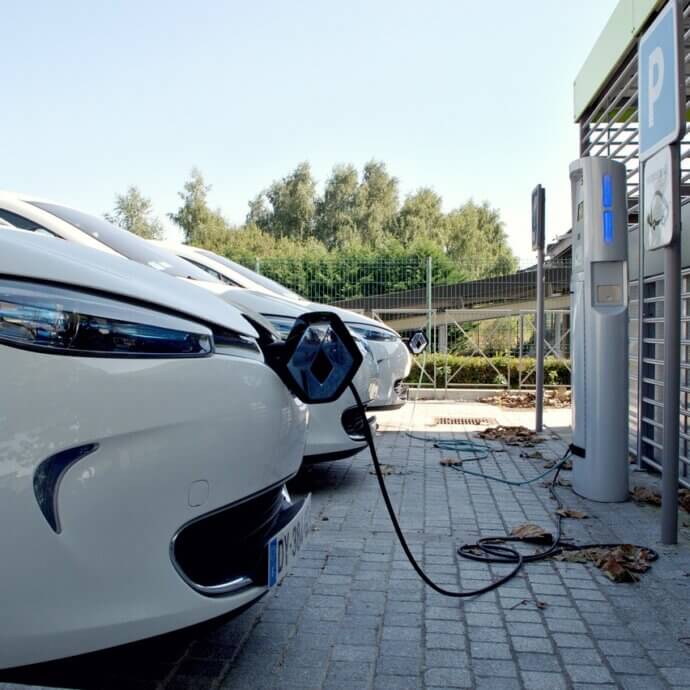 Place de stationnement. On aperçoit le devant d'un véhicule blanc électrique en plein chargement devant une borne de recharge, lors d'une journée d'été ensoleillée.